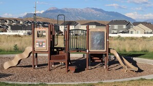 Ein Spielplatz mit Rutsche und Klettergerüst, umgeben von Kies und Rasen, mit Häusern und Bergen im Hintergrund.