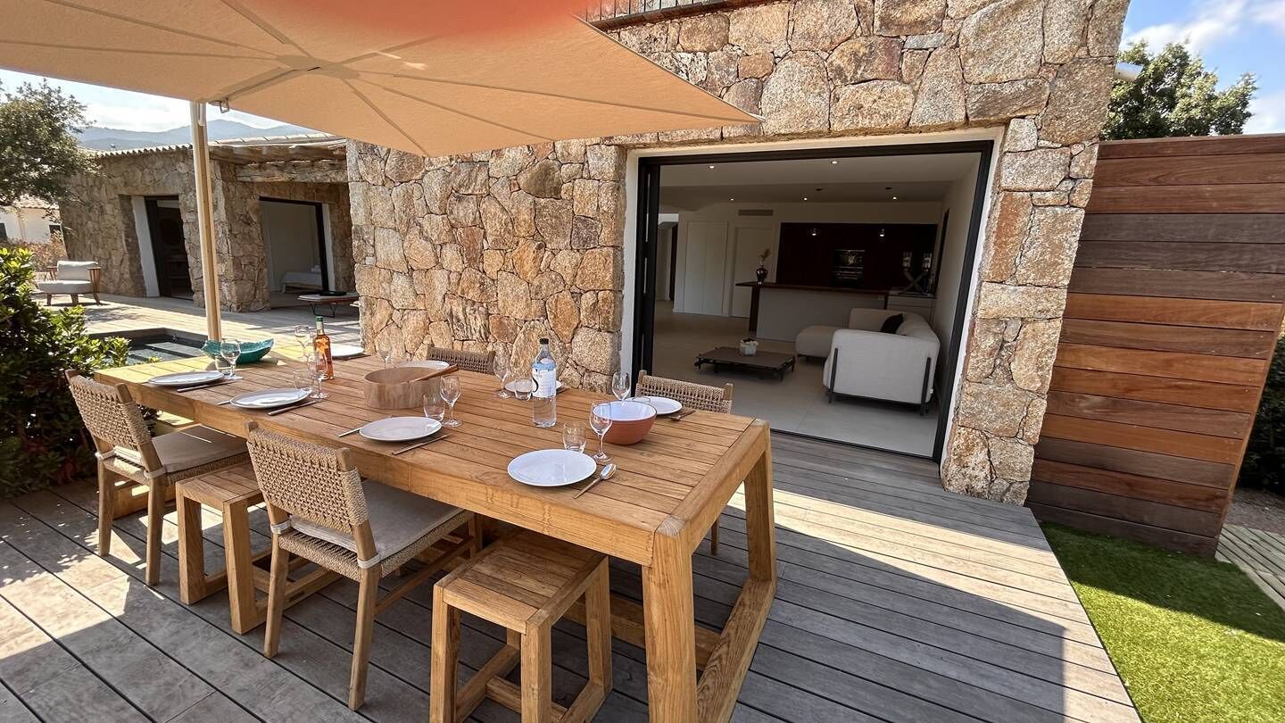 An outdoor wooden deck with a stone wall, a dining table set for four, and a covered seating area.