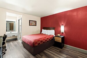 Habitación de hotel con cama, escritorio, silla, mesita de noche, lámpara y una pared con detalle en rojo.