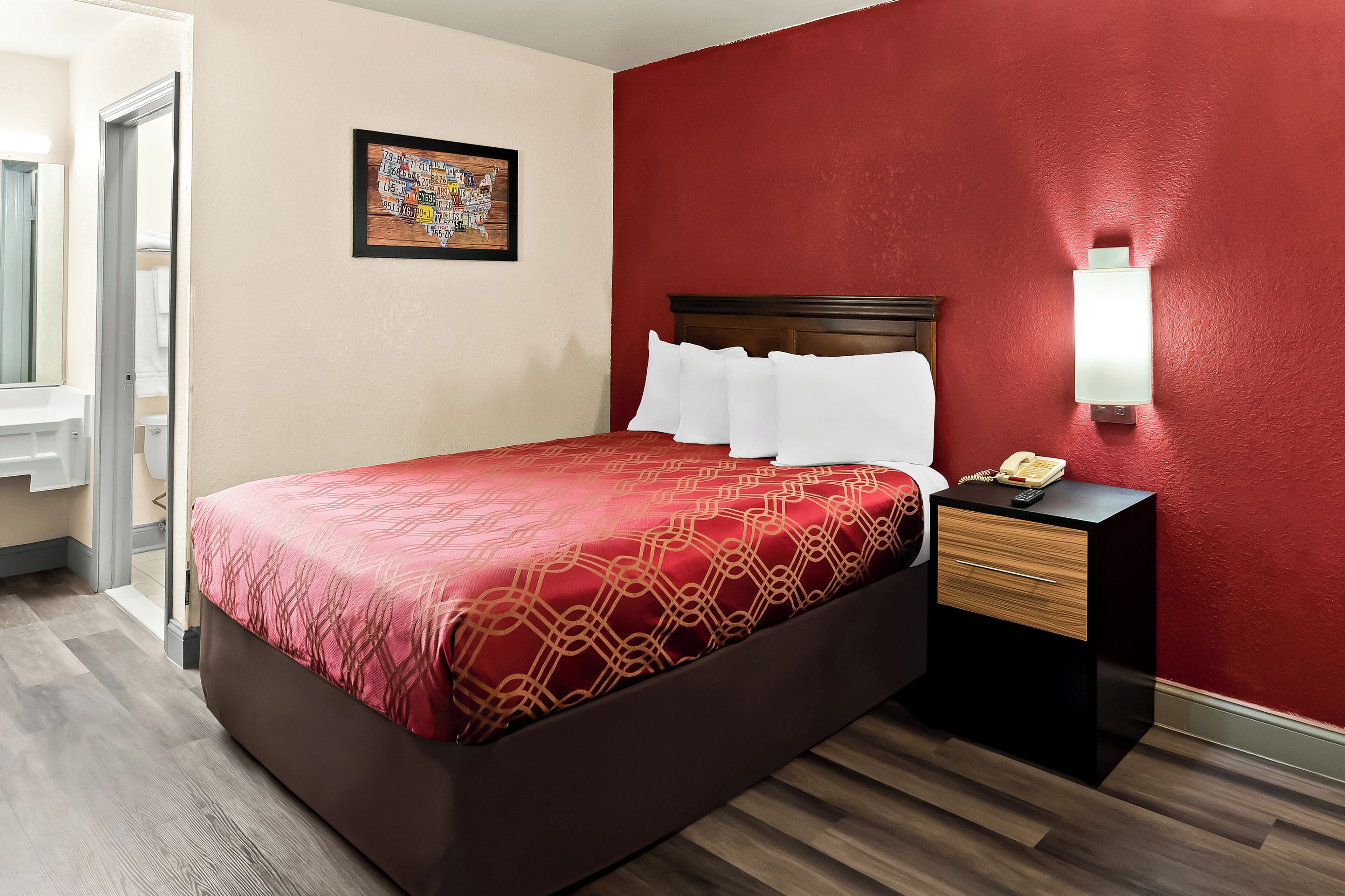 Habitación de hotel con cama, mesita de noche con lámpara, un cuadro en la pared y piso de madera.