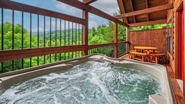 Ein Whirlpool mit einer Holzdielen-Terrasse und einem Tisch, umgeben von einem Holzzaun mit Blick auf grüne Hügel.