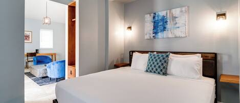 A bedroom with a large bed, a wooden headboard, a blue and white abstract painting, and a small table with a lamp.