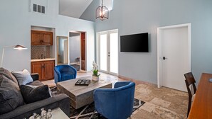 Een ruime woonkamer met een grote bank, een salontafel en blauwe stoelen. Er is een tv aan de muur bevestigd en een deur naar een andere kamer.