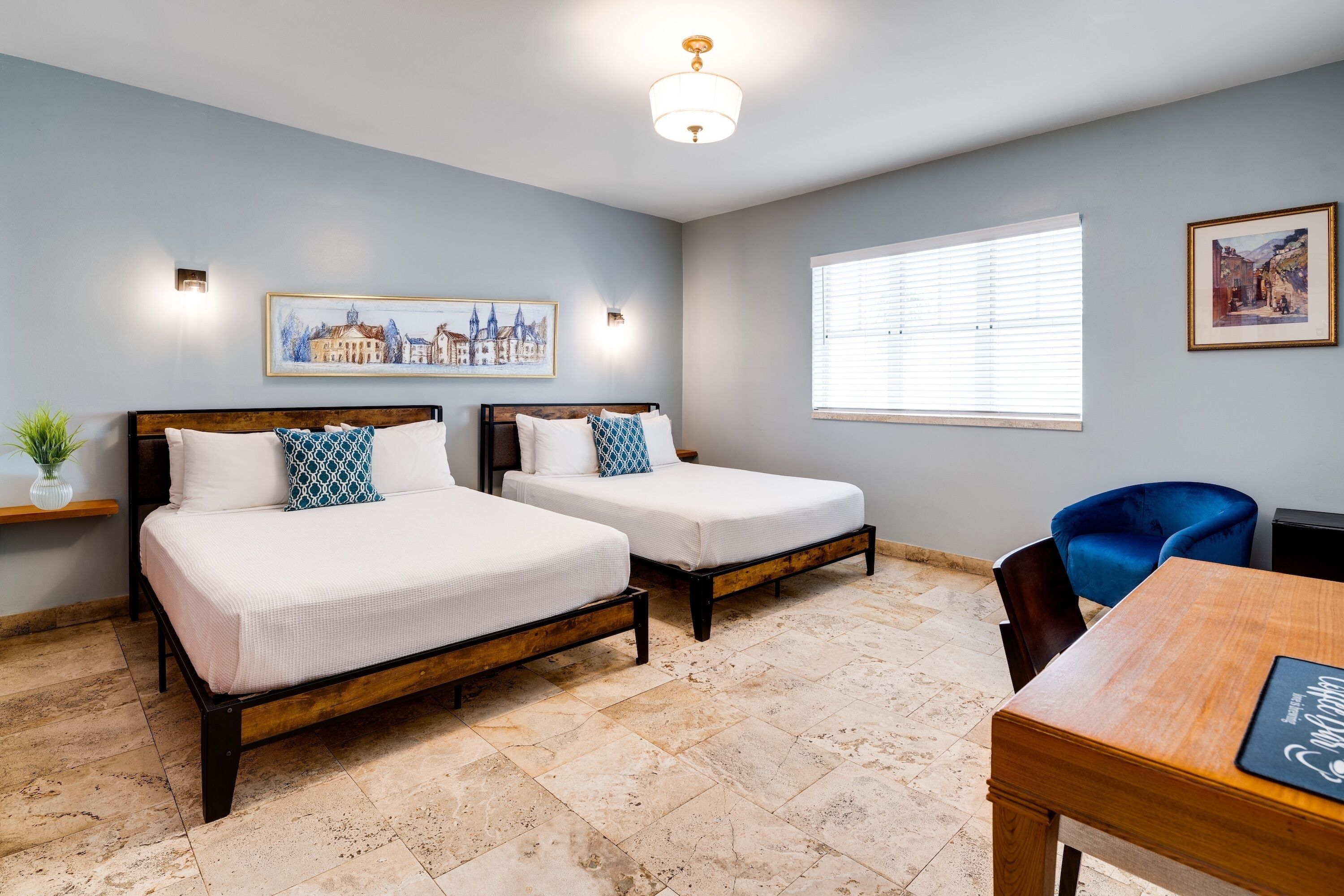 Une chambre d’hôtel avec deux lits, une table en bois et une fenêtre avec des stores.