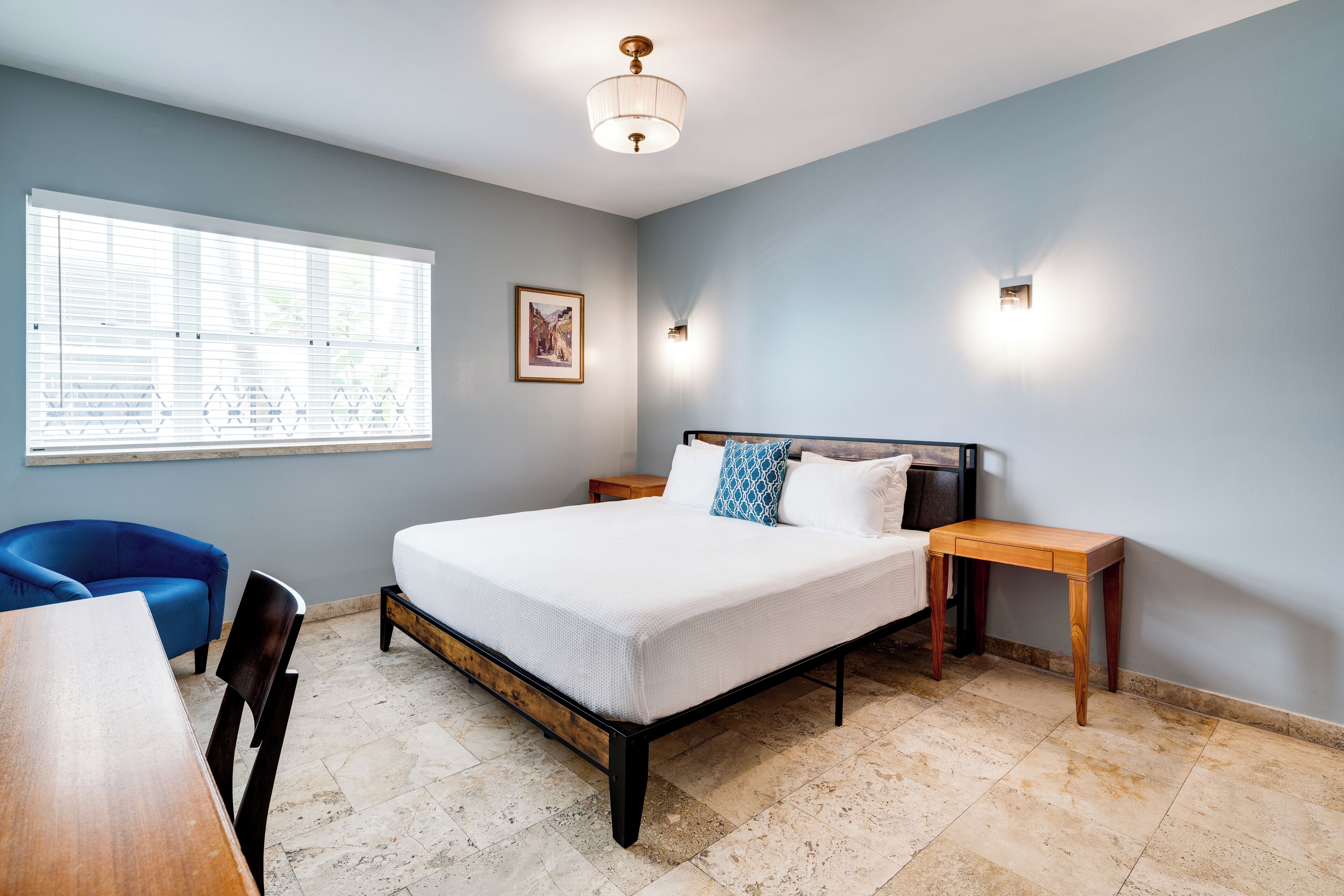 A bedroom with a bed, a desk, a chair, a window with blinds, and two wall-mounted lights.