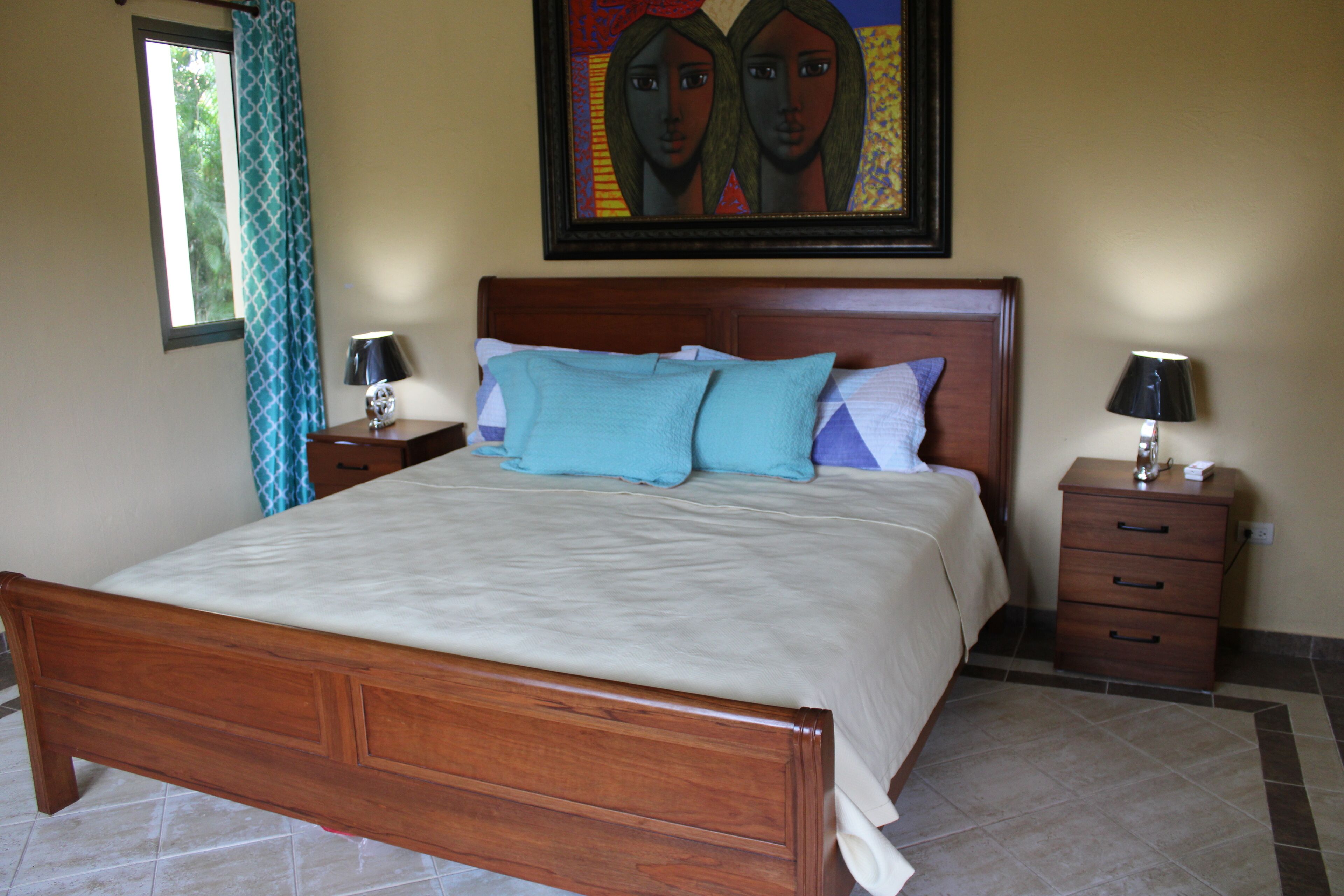 A bedroom with a wooden bed, two bedside tables with lamps, a window with a curtain, and a painting on the wall.