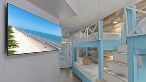 A room with a TV displaying a beach view, a bunk bed with a white pillow, and a staircase with a blue railing. - Dunes #205 | Remodeled Condo • Community Pool • 1 Min Walk to Beach (South Padre Island)