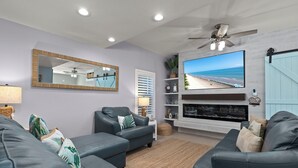 A modern living room with two grey leather sofas, a large flat-screen TV, a ceiling fan, and a beach view on the screen. - Dunes #205 | Remodeled Condo • Community Pool • 1 Min Walk to Beach (South Padre Island)