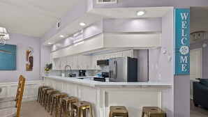 A modern kitchen with white cabinetry, a central island with stools, and a 'WELCOME' sign. - Dunes #205 | Remodeled Condo • Community Pool • 1 Min Walk to Beach (South Padre Island)