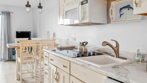 Een moderne keuken met een witte magnetron, een roestvrijstalen waterkoker en een wastafel met een gouden kraan. Er staan houten stoelen bij een kleine tafel, en aan de muur hangt een schilderij van een vis.