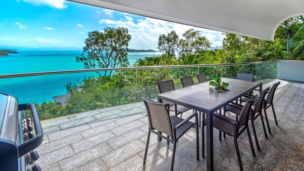 A balcony with a table and chairs, overlooking a coastal view with trees and the sea. - Waves 1 Hamilton Island - oceanviews, golf buggy and transfers (Hamilton Island)