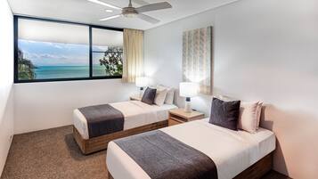 A hotel room with two beds, a ceiling fan, a large window with a view of the sea, and a painting on the wall.