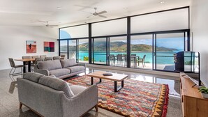 A modern living room with a gray sofa, a coffee table, and a colorful rug. There is a dining area with chairs and a table, and a large window offering a view of the sea and hills. - Waves 2 Hamilton Island - oceanviews, golf buggy and transfers (Hamilton Island)