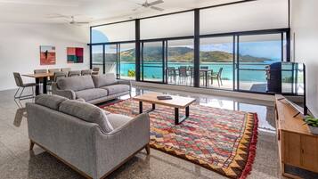 A modern living room with a gray sofa, a coffee table, and a colorful rug. There is a dining area with chairs and a table, and a large window offering a view of the sea and hills.