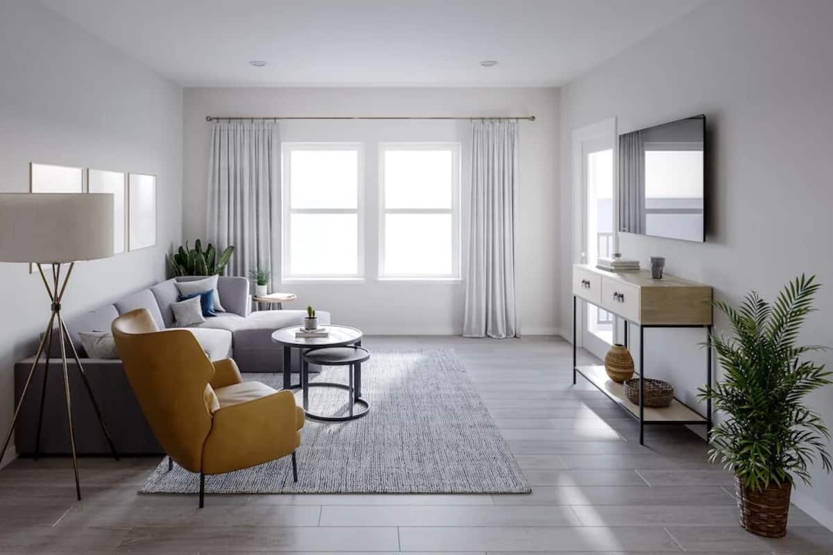 Un salon moderne comprenant un canapé, un fauteuil, une table basse et un lampadaire. De grandes fenêtres sont habillées de rideaux, et on y trouve également un téléviseur à écran plat ainsi qu’une petite table d’appoint agrémentée d’objets décoratifs.