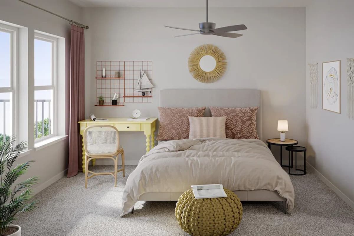 Une chambre à coucher comprenant un lit, un bureau, une chaise, un pouf rond, un ventilateur de plafond et une fenêtre avec des rideaux.