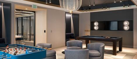 A modern lounge area with a foosball table, comfortable seating, a pool table, and a large TV screen.
