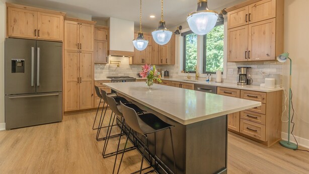 Een moderne keuken met houten kastjes, een roestvrijstalen koelkast, een centraal eiland met barkrukken, hanglampen en uitzicht op groen buiten.