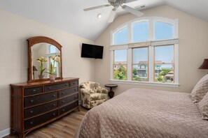 A bedroom with a large bed, a dresser, a chair, a TV, and a window with a view of houses.