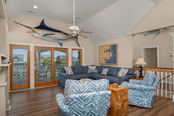 A spacious living room with a large sectional sofa, two patterned armchairs, and a wooden coffee table. A ceiling fan and a large fish decoration hang from the ceiling.
