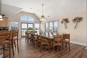 A spacious dining area with a large table and wooden chairs, a ceiling fan, and large windows offering views of the outdoors. - Southern Comfort: Semi-Oceanfront, Private Heated Pool, Elevator, Hot Tub, Dog Friendly! (Nags Head)