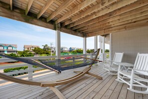 Una terraza de madera con una hamaca, mecedoras blancas y vistas a casas al fondo.