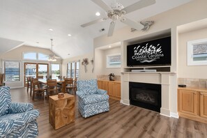 A spacious living room with a fireplace, a dining area, and a large TV screen displaying 'Coastal Carolina Vacations and Sales'.
