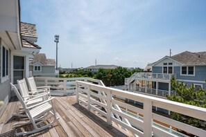 A wooden deck with white railings and rocking chairs, overlooking a residential area with houses and a streetlight. - Southern Comfort: Semi-Oceanfront, Private Heated Pool, Elevator, Hot Tub, Dog Friendly! (Nags Head)