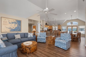 Uma sala de estar ampla, com um sofá azul, poltronas estampadas, uma mesa de centro de madeira e um espaço de jantar com cadeiras e mesa de madeira.