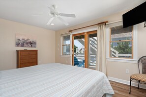 Un dormitorio con cama, ventilador de techo, cómoda, silla y ventana que da a una escalera y zona exterior.