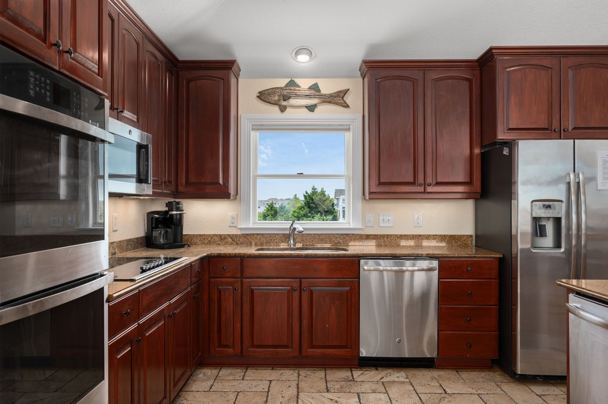 A kitchen with wooden cabinets, stainless steel appliances, a dishwasher, and a window with a view of the outside.