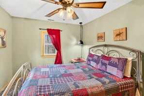 A bedroom with a metal bed frame, a quilted bedspread, a ceiling fan, a window with red curtains, and wall art.