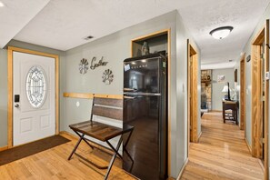 Un couloir avec un banc en bois, un réfrigérateur noir, une porte ornée d’un panneau de verre décoratif et un mur sur lequel on peut lire le mot « Gather » et sur lequel est accrochée une horloge.