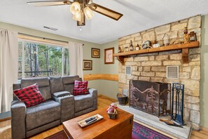 A living room with a stone fireplace, a sofa with red and black checkered pillows, a wooden coffee table, and a ceiling fan.