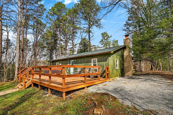 A cabin with a wooden deck, a chimney, and a gravel driveway surrounded by trees.