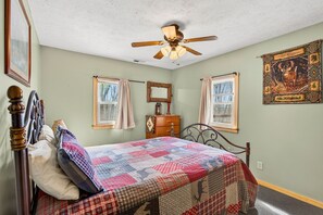 A bedroom with a bed, a ceiling fan, a dresser, a window with curtains, and a wall tapestry. - Lazy Lane Cabins - Bobcat Hideaway (South Bloomingville)