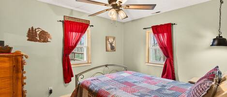 A bedroom with a bed, a ceiling fan, two windows with red curtains, a wall clock, and a wooden dresser.