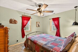 Une chambre à coucher avec un lit, un ventilateur de plafond, deux fenêtres avec des rideaux rouges, une horloge murale et une commode en bois.