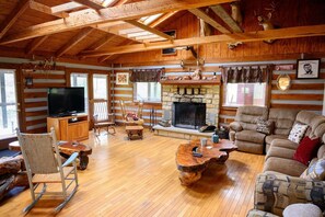 A log cabin interior with a stone fireplace, wooden furniture, and a television.