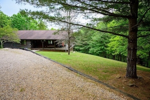 A cabin with a gravel driveway, surrounded by trees and a grassy slope.
