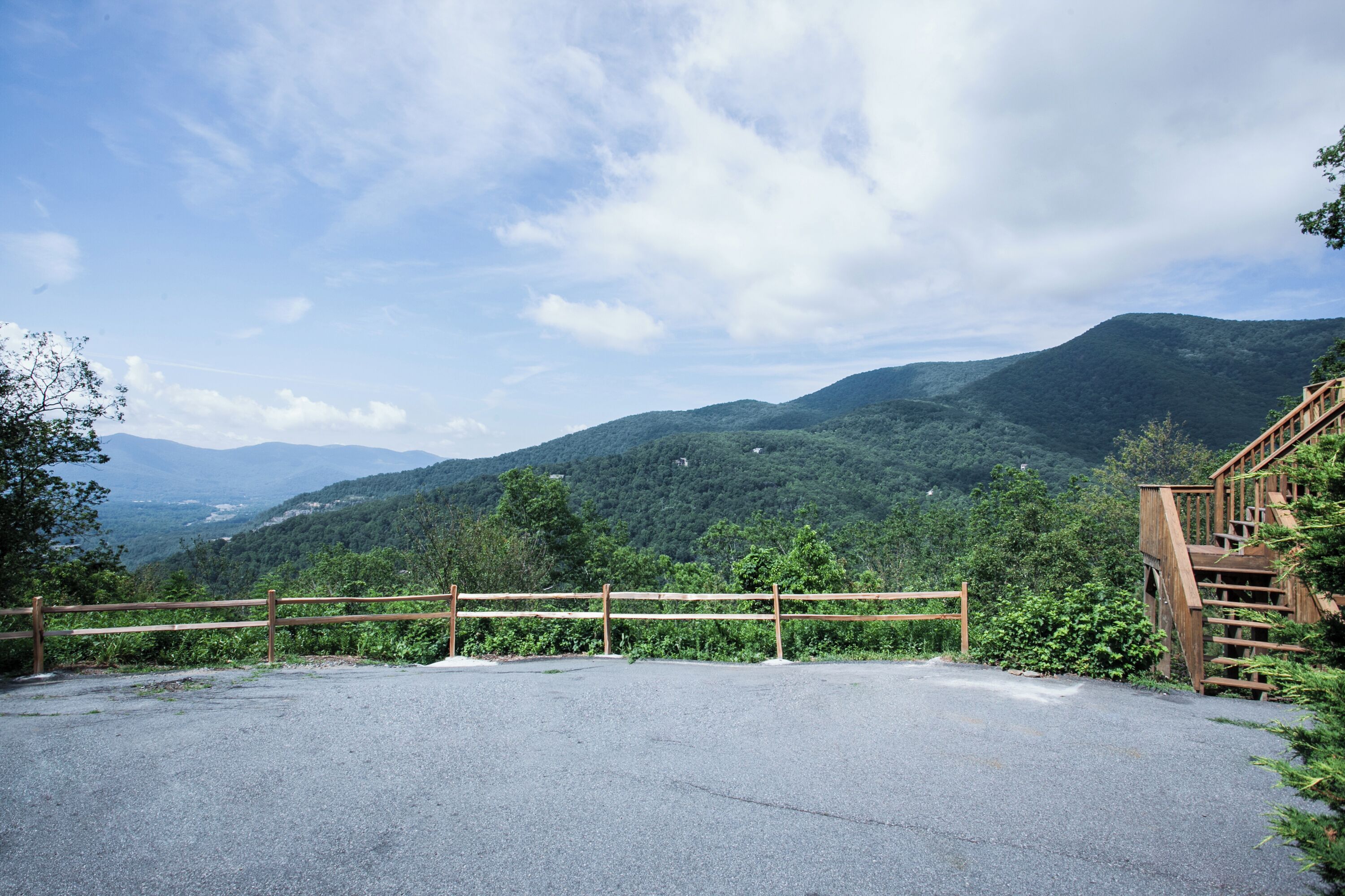 A paved parking area with a wooden fence and staircase leading to a viewing platform overlooking a mountainous landscape.