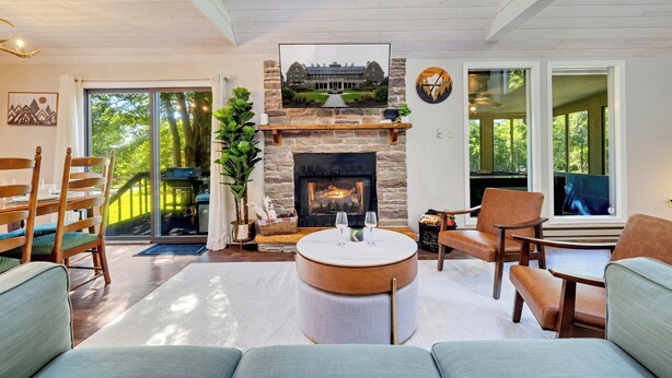A modern living room with a stone fireplace, comfortable seating, and a large window offering a view of the outdoors. - New! Hot Tub, Fire-pit, Shuffleboard, Games Room, Kids Area in Gated Community (Albrightsville)