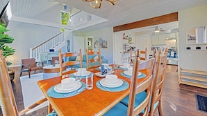 A dining area with a wooden table set for four, surrounded by chairs. A staircase leads to an upper level, and a kitchen is visible in the background. - New! Hot Tub, Fire-pit, Shuffleboard, Games Room, Kids Area in Gated Community (Albrightsville)