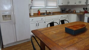 Une cuisine avec des meubles blancs, une table en bois et une fenêtre avec des stores.