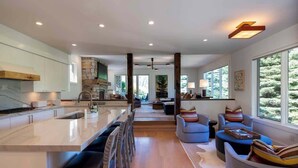 A modern kitchen with a central island, a dining area with chairs, a living room with sofas, and large windows offering a view of trees. - Abode at Sunny Knob Hill, scenic setting, hot tub (Ketchum)