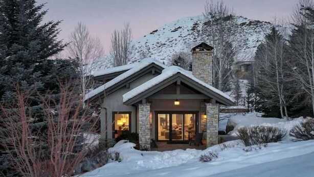 A snow-covered mountain house with a stone chimney, a lit interior, and a snow-covered driveway. - Abode at Sunny Knob Hill, scenic setting, hot tub (Ketchum)