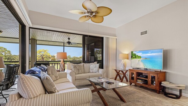 A spacious living room with a large TV, comfortable seating, and a view of the outdoors. - Bonita Bay Condo, Beach Club Included! Corner unit! (Bonita Springs)