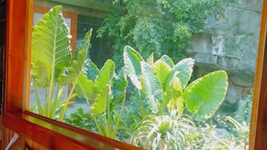 Una habitación con una cama de madera, un ventanal con techo de paja y abundante vegetación afuera.