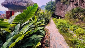 Un sendero de piedra conduce a una construcción rústica con techo de paja, rodeada de exuberante vegetación y una ladera rocosa.