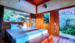 A bedroom with a bed, wooden walls, a stone wall, and a view of greenery.